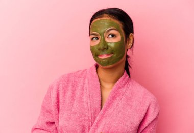 Genç Venezuelalı kadın, pembe arka planda izole edilmiş bornoz ve yüz maskesi takıyor. Kafası karışık ve kararsız..