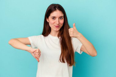 Mavi arka planda izole edilmiş genç beyaz kadın baş parmaklarını aşağı ve yukarı kaldırıyor, zor seçim konsepti