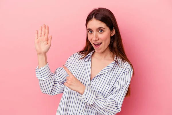 Pembe arka planda izole edilmiş genç beyaz kadın gülümseyerek beş numarayı parmaklarıyla gösteriyor..