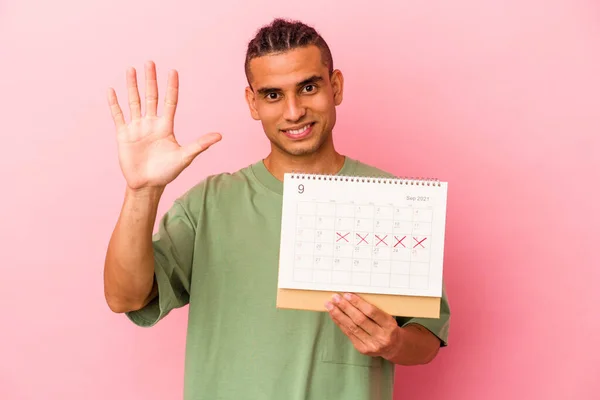 若いですVenezuelan男保持Aカレンダー隔離上のピンクの背景笑顔陽気に示す番号5とともに指 — ストック写真
