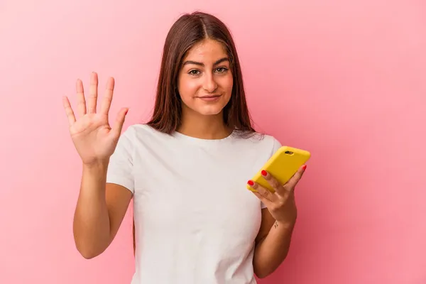 年轻的高加索女人拿着一部粉色背景的手机 高兴地微笑着 用手指展示着5号 — 图库照片