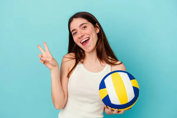 青を背景に孤立したバレーボールをしている若い白人女性喜びと屈託のない指で平和のシンボルを示す — ストック写真