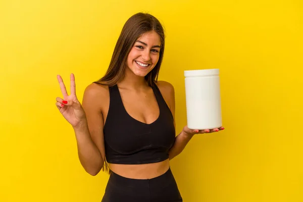 黄色の背景に単離されたタンパク質ボトルを保持若い白人女性喜びと屈託のない指で平和のシンボルを示す — ストック写真
