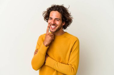 Beyaz arka planda izole olmuş genç beyaz bir adam gülümsüyor ve kendinden emin, çenesine elleriyle dokunuyor..