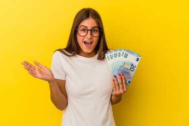 Sarı arka planda izole edilmiş banknotları tutan genç beyaz kadın şaşırdı ve şok oldu..