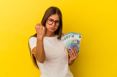 Sarı arka planda izole edilmiş banknotlar tutan genç beyaz kadın kameraya yumruk gösteriyor, agresif yüz ifadesi var..