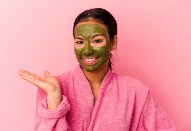 Genç Venezuelalı kadın, pembe arka planda izole edilmiş bornoz ve yüz maskesi takıyor. Bir avuç içinde fotokopi alanı var ve belinde başka bir el tutuyor..