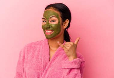 Bornoz ve yüz maskesi takan genç Venezuelalı kadın pembe arka planda izole edilmiş, parmaklarıyla bir cep telefonu hareketi gösteriyor..