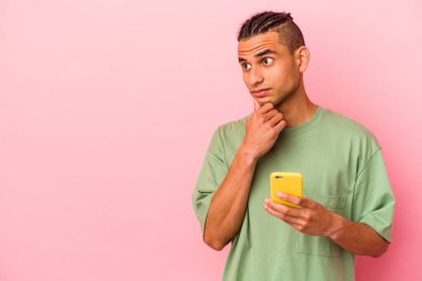Genç Venezuelalı bir adam pembe arka planda izole edilmiş bir cep telefonu tutuyor şüpheli ve şüpheci bir ifadeyle yana bakıyor..
