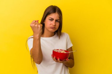 Sarı arka planda izole edilmiş mısır gevreği tutan genç beyaz kadın kameraya yumruk ve agresif yüz ifadesi gösteriyor..