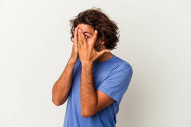 Beyaz arka planda izole edilmiş genç beyaz bir adam parmaklarının arasından korkmuş ve gergin bir şekilde göz kırpıyor..