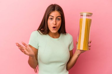 Elinde makarna kavanozu tutan beyaz bir kadın pembe arka planda izole edilmiş, şaşırmış ve şok olmuş..