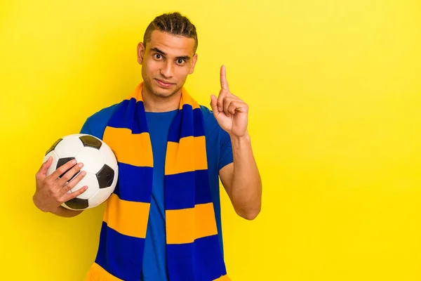 Młody Wenezuelczyk Ogląda Piłkę Nożną Odizolowaną Żółtym Tle Pokazuje Numer — Zdjęcie stockowe
