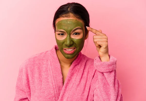 Venezuelalı genç bir kadın pembe arka planda izole edilmiş bornoz ve yüz maskesi giyiyor işaret parmağıyla yaptığı hayal kırıklığı jestini gösteriyor..