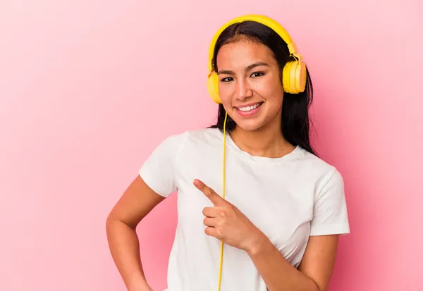 若いベネズエラの女性はピンクの背景に孤立した音楽を聞いて笑顔と脇を指して 空白のスペースで何かを示しています — ストック写真