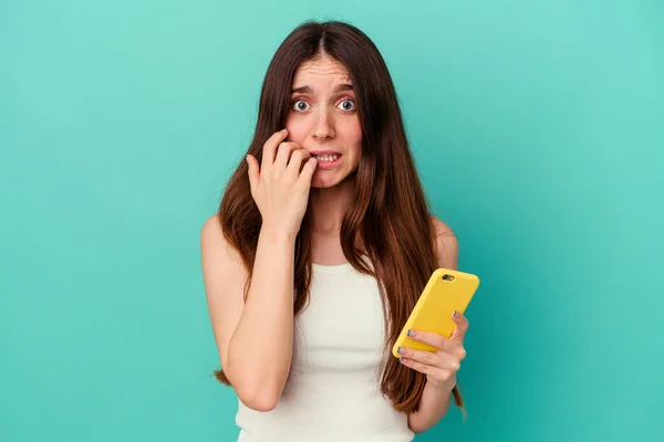 Jovem Caucasiana Segurando Telefone Celular Isolado Fundo Azul Mordendo Unhas — Fotografia de Stock