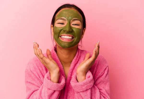 Genç Venezuelalı kadın pembe arka planda izole edilmiş bornoz ve yüz maskesi takıyor. Hoş bir sürprizle karşılaşıyor, heyecanlı ve el kaldırıyor..