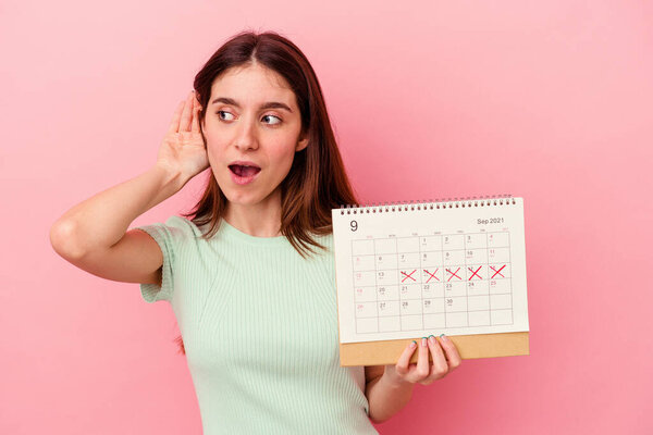 Young caucasian woman holding a calendar isolated on pink background trying to listening a gossip.