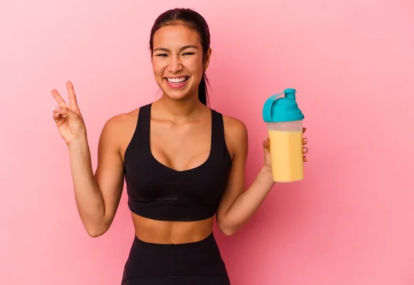 ピンクの背景に単離されたタンパク質シェイクを飲む若いベネズエラの女性喜びと屈託のない指で平和のシンボルを示す — ストック写真