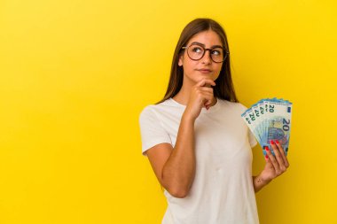 Sarı arka planda izole edilmiş banknotları tutan genç beyaz kadın şüpheli ve şüpheci bir ifadeyle yana bakıyor..