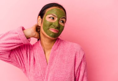 Genç Venezuelalı kadın, pembe arka planda izole edilmiş bornoz ve yüz maskesi takıyor. Kafasının arkasına dokunuyor, düşünüyor ve bir seçim yapıyor..