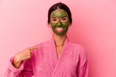Bornoz ve yüz maskesi takan genç beyaz bir kadın pembe arka planda izole edilmiş ve gömlek fotokopi alanını işaret ediyor, gururlu ve kendinden emin.