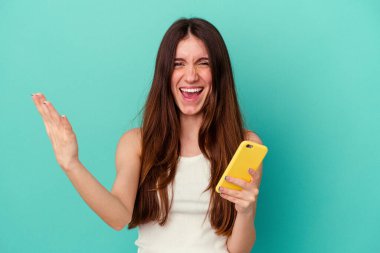 Mavi arka planda cep telefonu tutan beyaz bir kadın hoş bir sürpriz yaşıyor, heyecanlı ve el kaldırıyor..