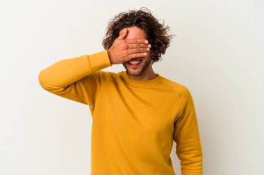 Beyaz arka planda soyutlanmış beyaz tenli genç bir adam elleriyle gözlerini kapatıyor, geniş bir gülümsemeyle bir sürpriz bekliyor..