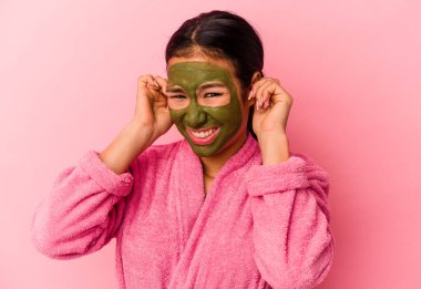 Bornoz ve yüz maskesi takan genç Venezuelalı kadın pembe arka planda izole edilmiş kulakları elleriyle kapatıyor..