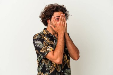 Beyaz arka planda izole edilmiş genç beyaz bir adam parmaklarının arasından korkmuş ve gergin bir şekilde göz kırpıyor..