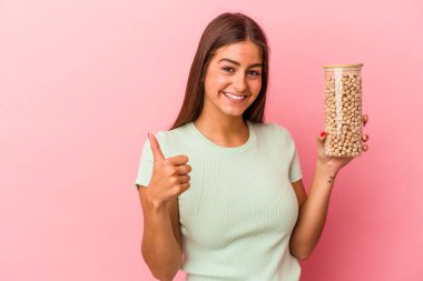 Pembe arka planda izole edilmiş nohut kavanozunu tutan beyaz bir kadın gülümsüyor ve başparmağını kaldırıyor.