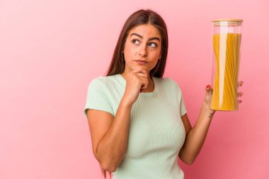 Pembe arka planda makarna kavanozu tutan beyaz bir kadın şüpheli ve şüpheci bir ifadeyle yan yana bakıyor..