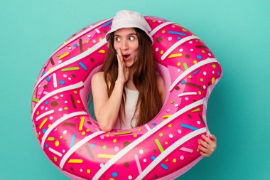 Mavi arka planda izole edilmiş şişme donutu olan genç beyaz kadın gizli bir sıcak fren haberi veriyor ve yan bakıyor.
