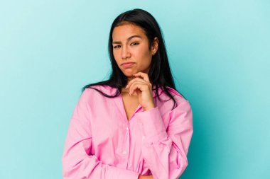 Genç Venezuelalı kadın mavi arka planda izole edilmiş düşünüyor ve yukarı bakıyor, yansıtıyor, düşünüyor, hayal kuruyor..