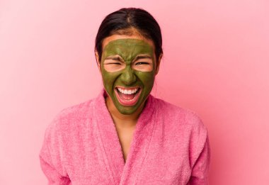 Genç Venezuelalı kadın, pembe arka planda izole edilmiş bornoz ve yüz maskesi takıyor. Çok sinirli ve agresif bağırıyor..