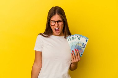 Sarı arka planda izole edilmiş banknotları tutan genç beyaz kadın çok sinirli ve agresif çığlıklar atıyor..