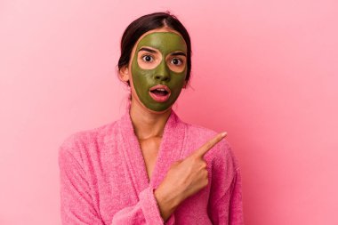 Pembe arka plana izole edilmiş bornoz ve yüz maskesi takan beyaz bir kadın yan tarafı işaret ediyor.