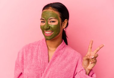 Genç Venezuelalı kadın pembe arka planda izole edilmiş bornoz ve yüz maskesi takıyor neşeli ve kaygısız parmaklarıyla barış sembolü gösteriyor..