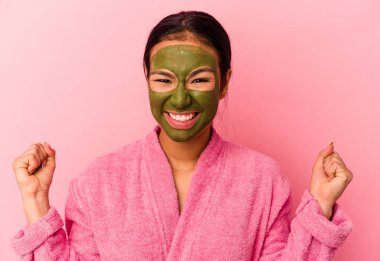 Genç Venezuelalı kadın bornoz ve yüz maskesi takıyor. Pembe arka planda izole edilmiş. Zaferden sonra yumruğunu kaldırıyor..