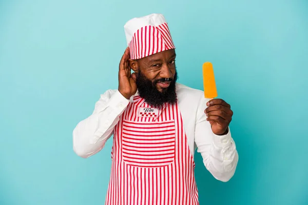 African american ice cream maker man holding an ice cream isolated on blue background trying to listening a gossip.