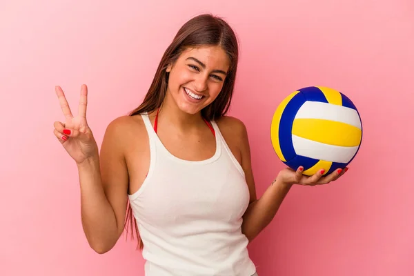 年轻的高加索女人拿着一个被粉色背景隔开的排球球 快乐而无忧无虑 用手指展示一个和平的象征 — 图库照片