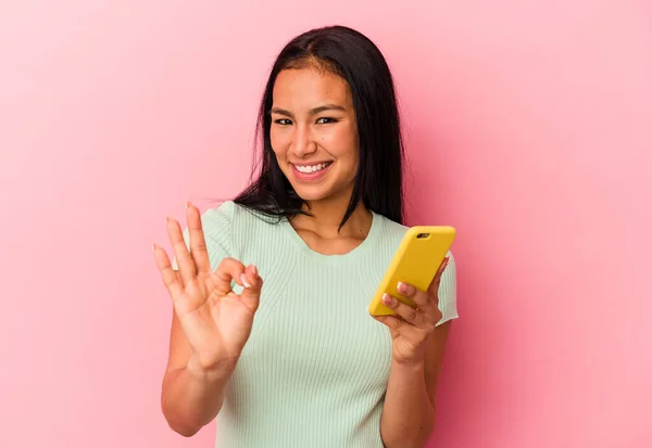 若いですベネズエラの女性を保持しています携帯電話ピンクの背景に孤立陽気で自信を持ってショーOkジェスチャー — ストック写真