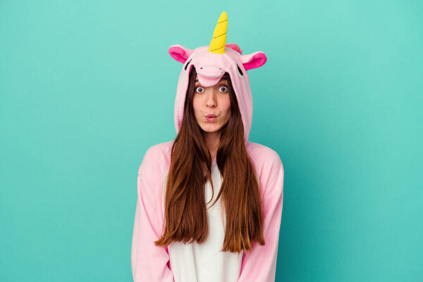 Young caucasian woman wearing a unicorn pajama isolated on blue background shrugs shoulders and open eyes confused.