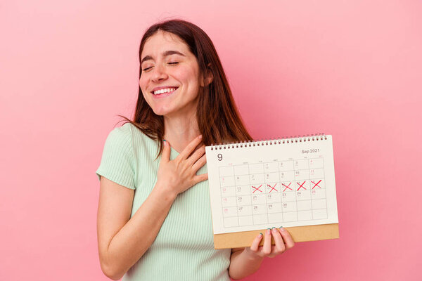 Молодая белая женщина, держащая календарь на розовом фоне, громко смеется, держа руку на груди..