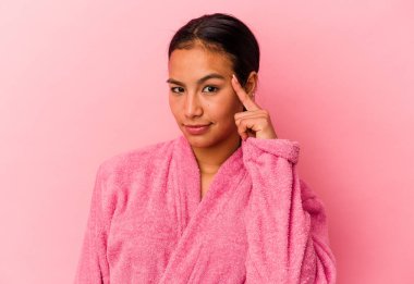 Genç Venezuelalı bir kadın pembe arka planda izole edilmiş bir bornoz giyiyor. Parmağıyla tapınağı işaret ediyor, düşünüyor, bir göreve odaklanıyor..