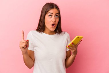 Elinde cep telefonu olan beyaz bir kadın pembe arka planda izole edilmiş ve ağzı açık bir şekilde üst tarafı işaret ediyor..