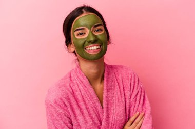 Pembe arka plana izole edilmiş bornoz ve yüz maskesi takan beyaz bir kadın gülüyor ve eğleniyor..