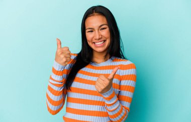 Genç Venezuelalı bir kadın mavi arka planda izole edilmiş. Baş parmağını kaldırıyor, gülümsüyor ve kendinden emin..