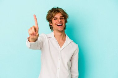 Makyajlı genç beyaz adam mavi arka planda bir numarayı parmağıyla gösteriyor..