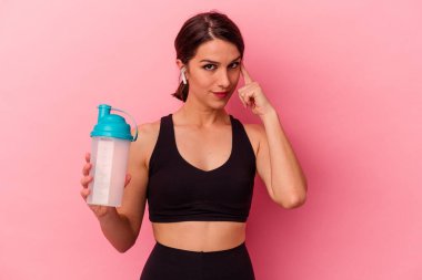 Beyaz bir kadın pembe arka planda izole edilmiş protein içeceği içiyor. Şakağı işaret ediyor, düşünüyor, bir göreve odaklanıyor..